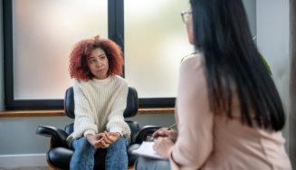 A young person with curly hair sits in a chair, engaged in a conversation with someone out of frame, in a warm, inviting room.